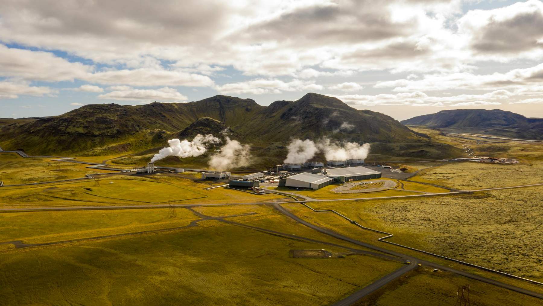 Steam rises from a geothermal power plant sits in a grassy field.