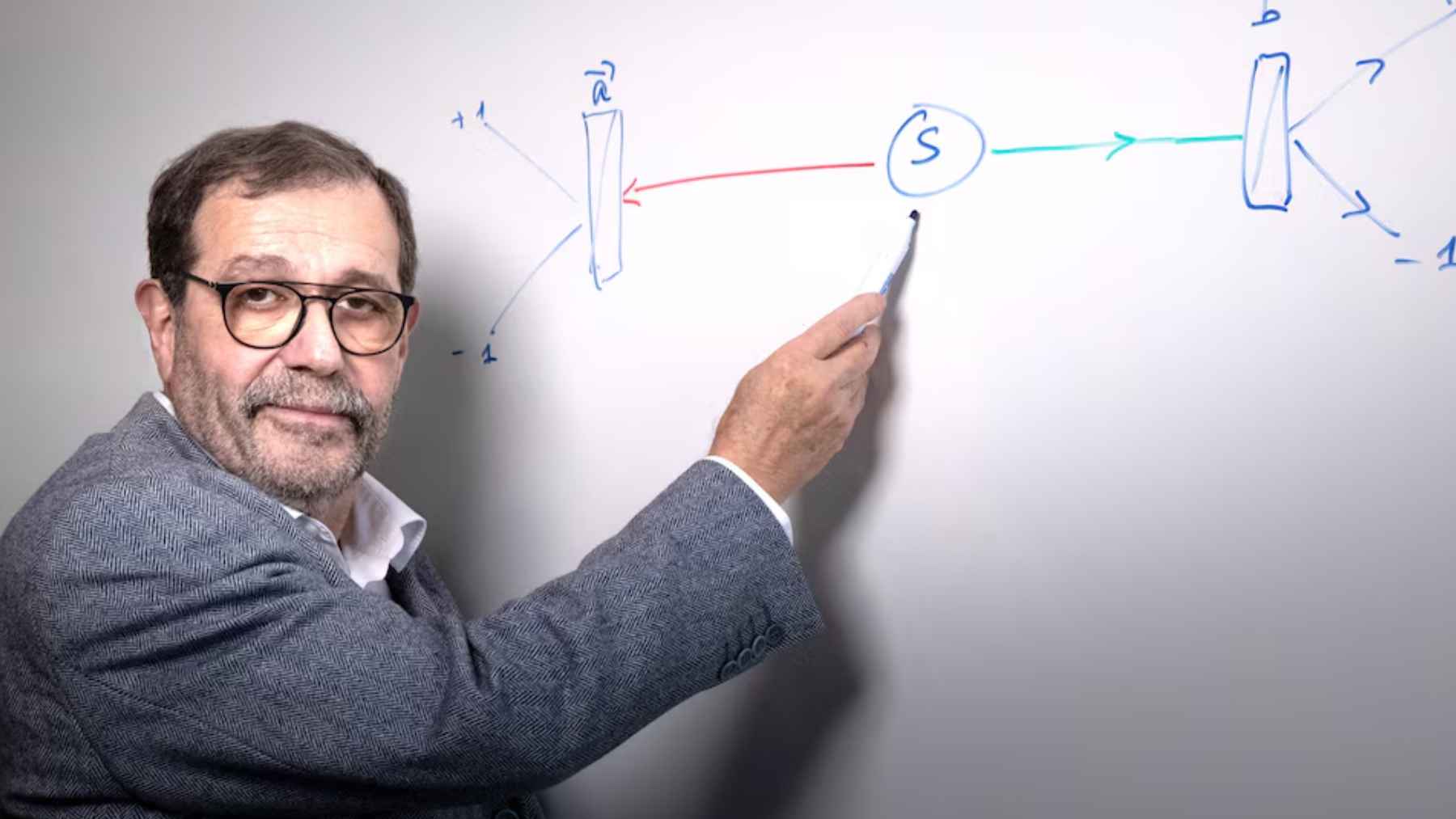 Alain Aspect señalando un esquema en una pizarra durante una explicación de física cuántica
