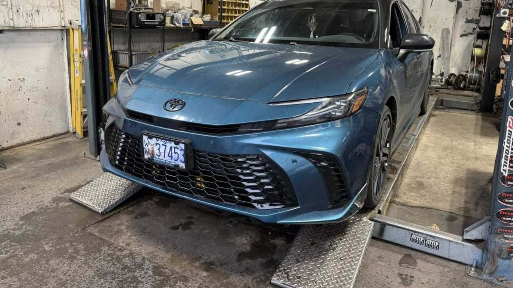 Toyota Camryin Guard Metallic 2023 en un elevador hidráulico en un taller de reparación, vista frontal de tres cuartos que muestra la carrocería completa y las herramientas circundantes.