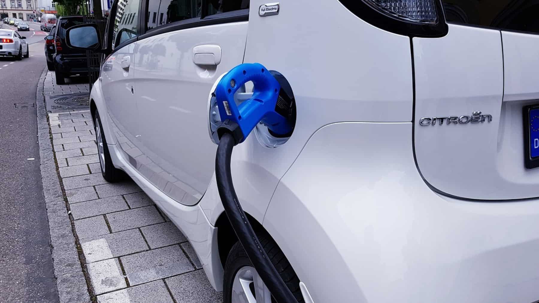 Coche eléctrico blanco cargando en la calle con un conector azul enchufado y el cable conectado al puerto de carga trasero.