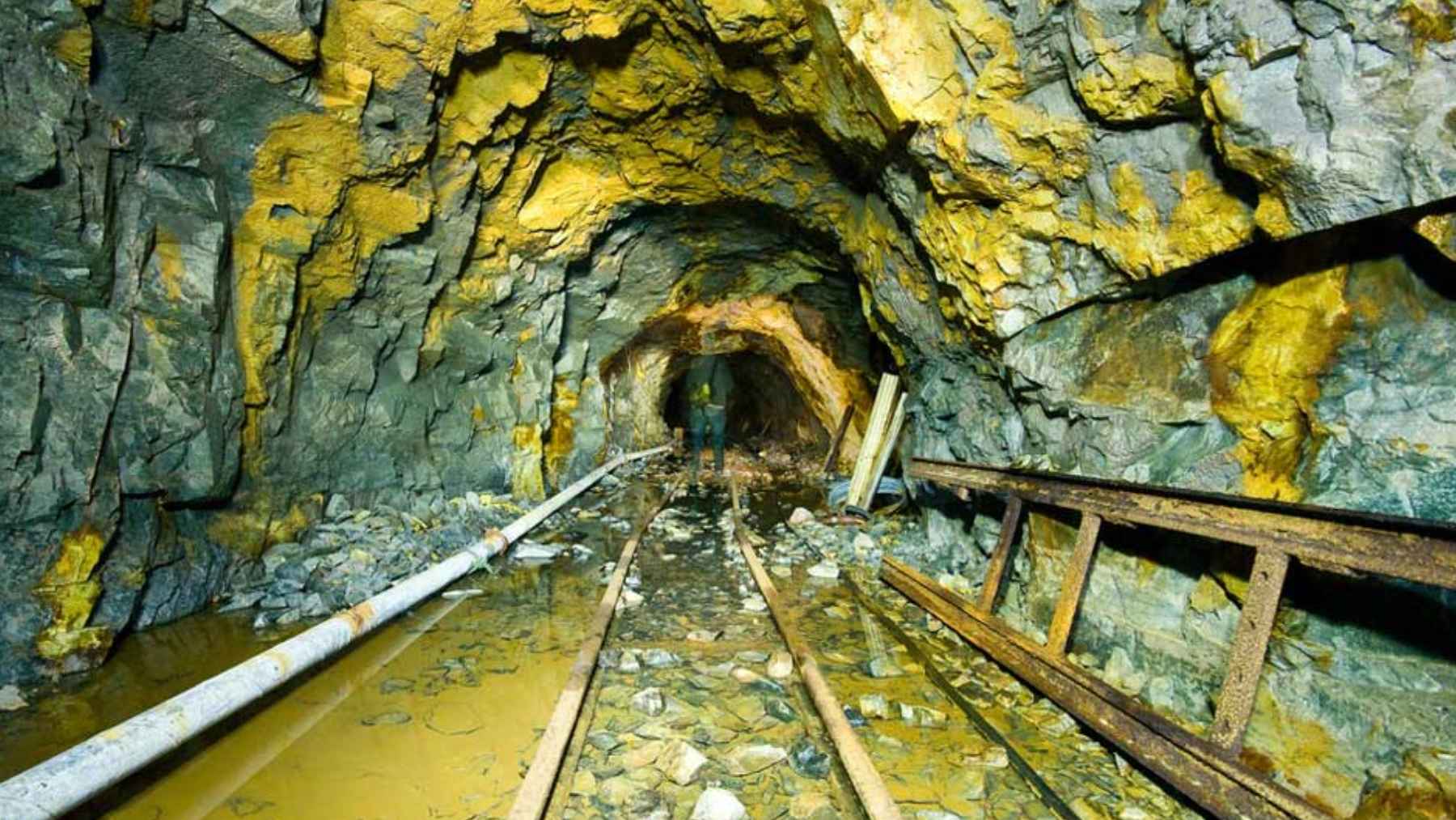 Galería de mina subterránea con vías y agua acumulada, asociada a la extracción de oro a gran profundidad